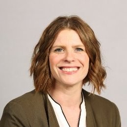 A woman with shoulder length light brown hair and wearing a brown jacket smiling at the camera.